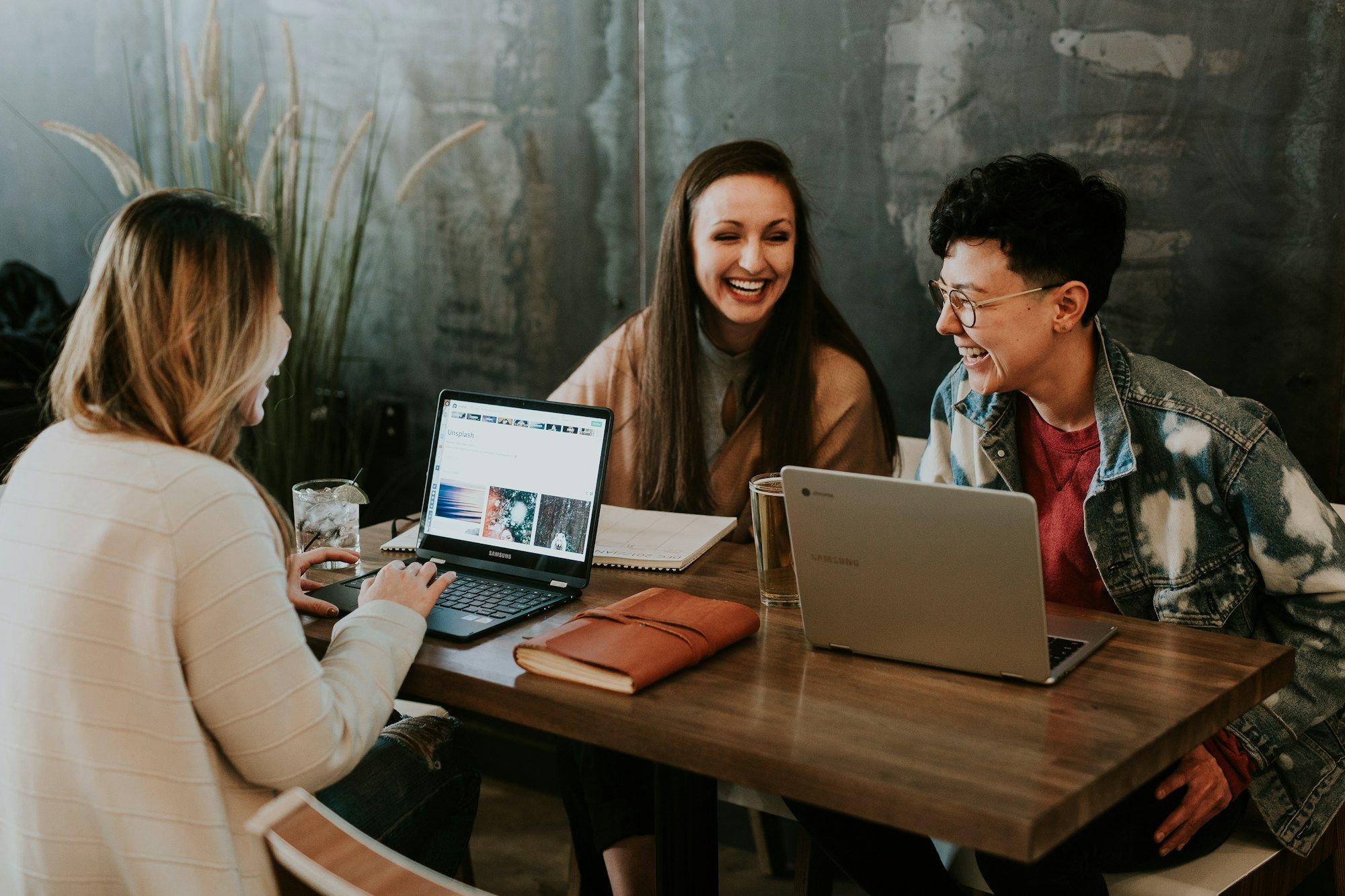 Three colleagues laughing together while working on laptops at a wooden table