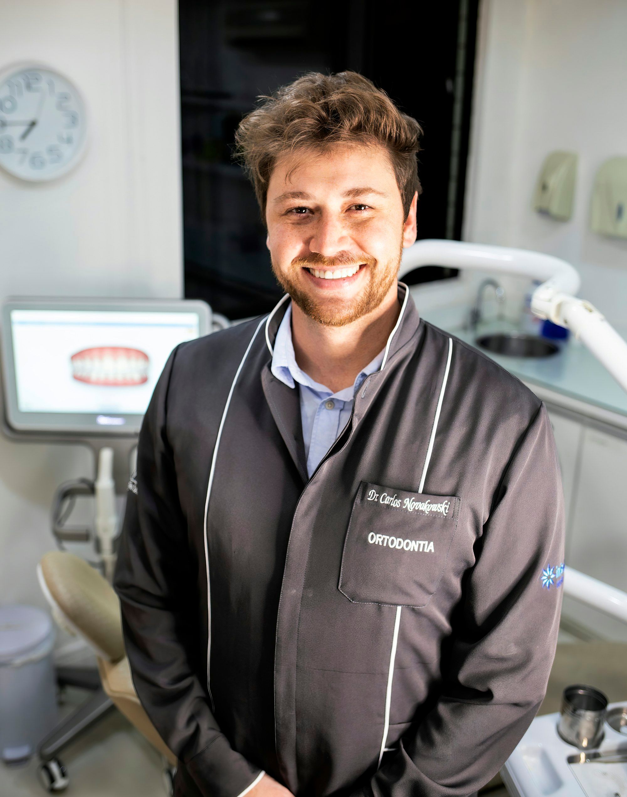 Smiling dentist in a dark clinic jacket standing in his treatment room