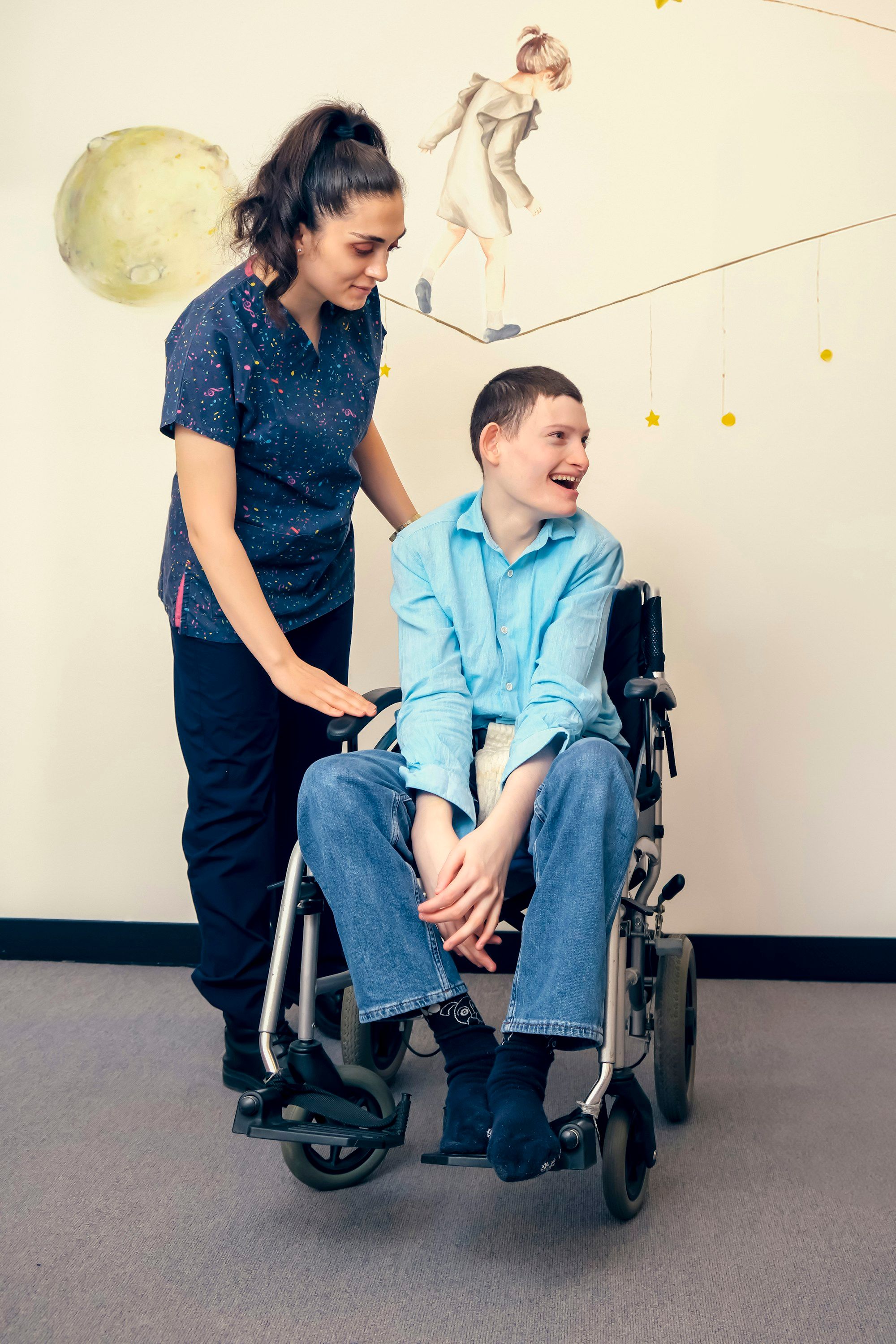 Support worker walking beside a smiling young man in a wheelchair in a bright corridor
