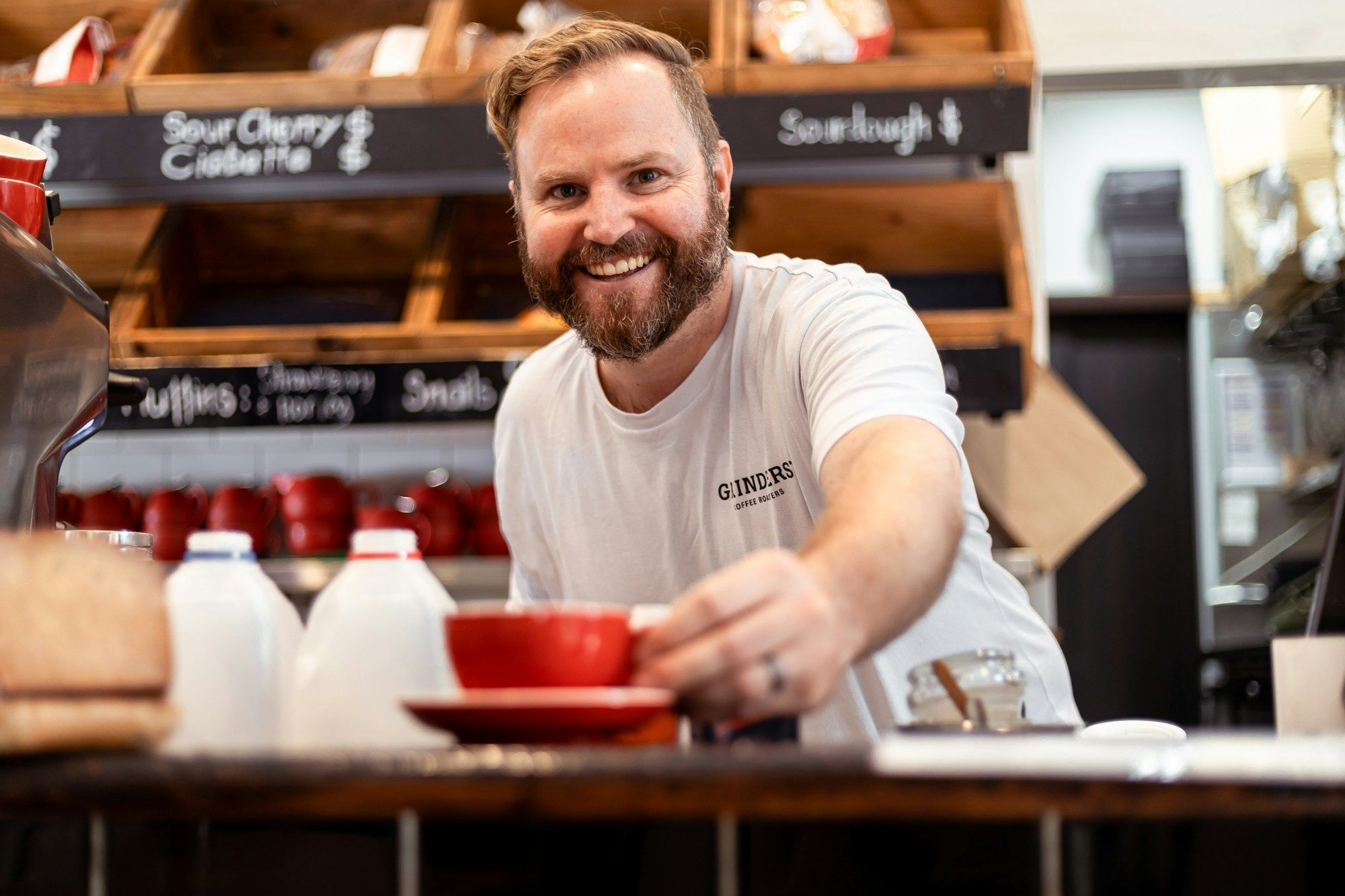 Smiling café owner handing a red cup of coffee across the counter of his bakery café